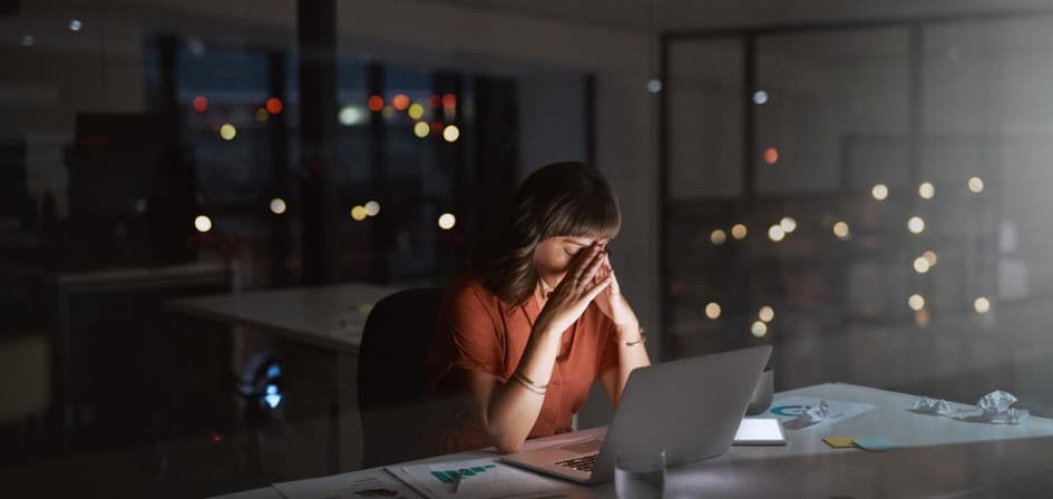 How to Deal with Workaholism. An image of a a young business woman looking stressed out while working on a laptop in an office at night.