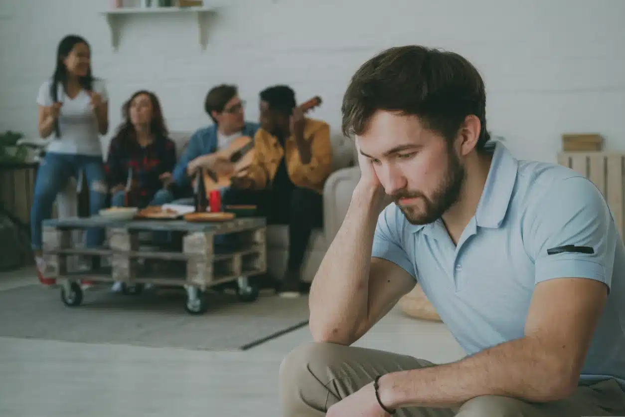 An image of a man displaying anhedonia symptoms whilst hanging out with friends. The group of friends can be seen in the background having fun and playing the guitar, whilst a male individual sits apart from the rest of the group, looking down.