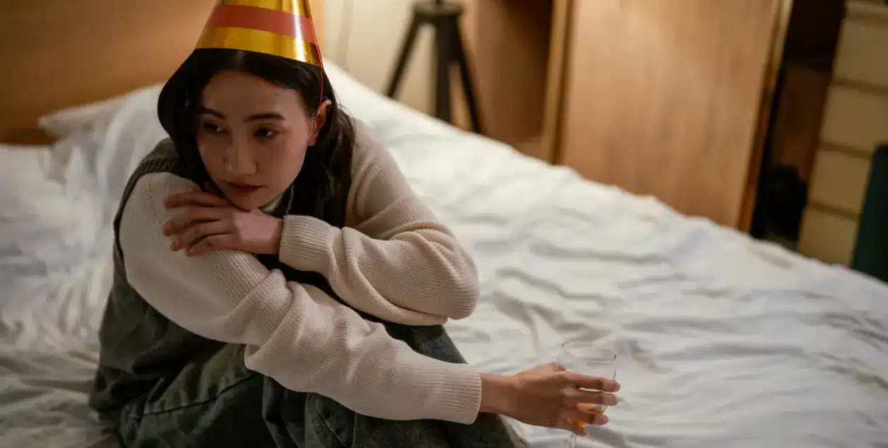 How Do I Know if I’m an Alcoholic? A woman looking deep in thought whilst sat on a bed holding a glass with a party hat on.